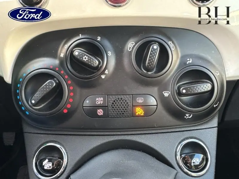 Gros plan sur les commandes de climatisation et ventilation d'une Fiat 500 blanche, avec réglages manuels et voyants allumés.