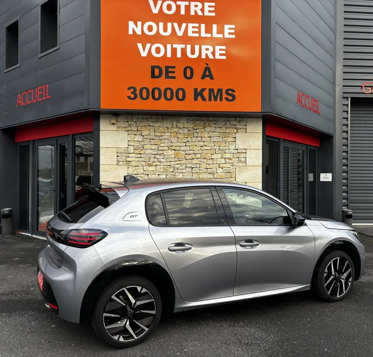 Peugeot 208 Hybrid 110 e-DCS6 GT gris clair vue de profil côté droit devant un bâtiment avec panneau orange "votre nouvelle voiture".