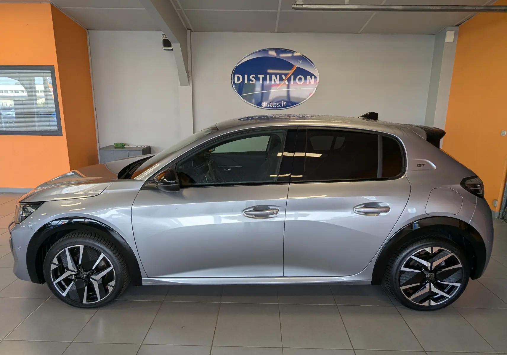 Profil côté gauche d'une Peugeot 208 HYBRID 145CH GT gris Artens avec jantes bi-ton et vitres teintées en showroom.