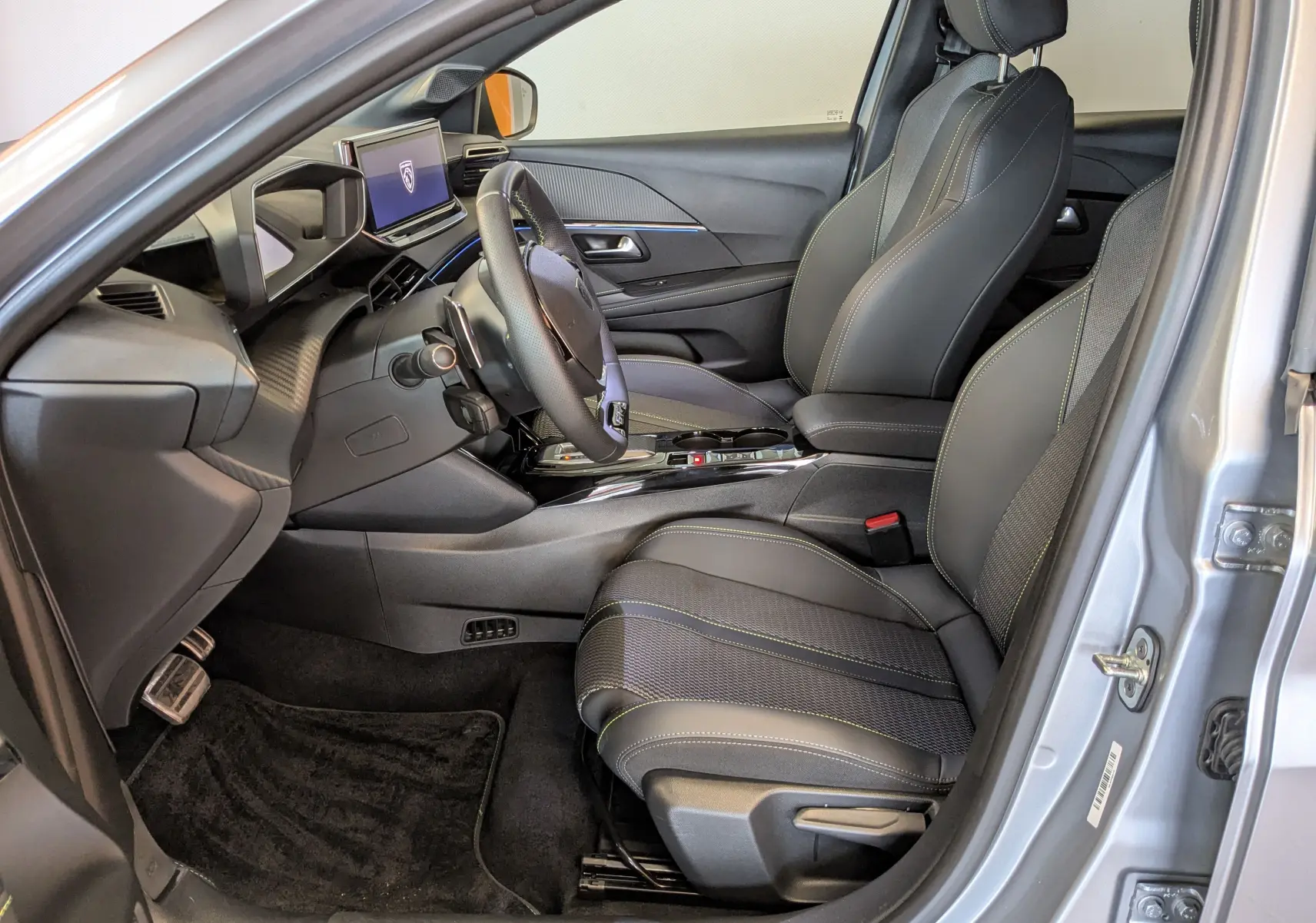 Vue intérieure côté conducteur de la Peugeot 208 Hybrid 145ch gris Artens, sièges noirs avec surpiqûres vertes et tableau de bord moderne.