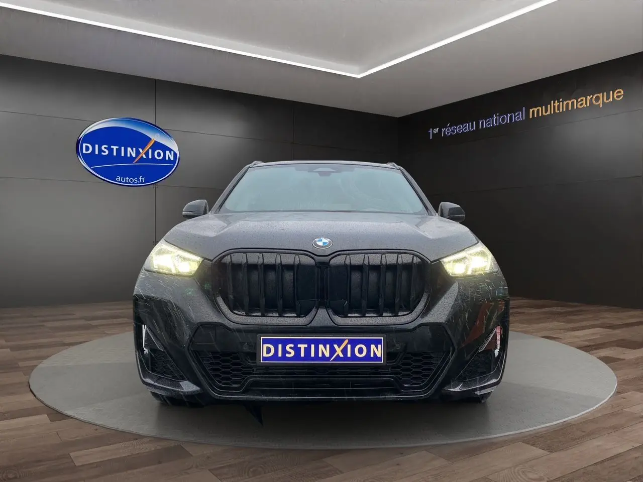 Vue frontale d'un BMW X1 XDRIVE 20D M SPORT noir Saphir avec phares allumés dans un showroom.
