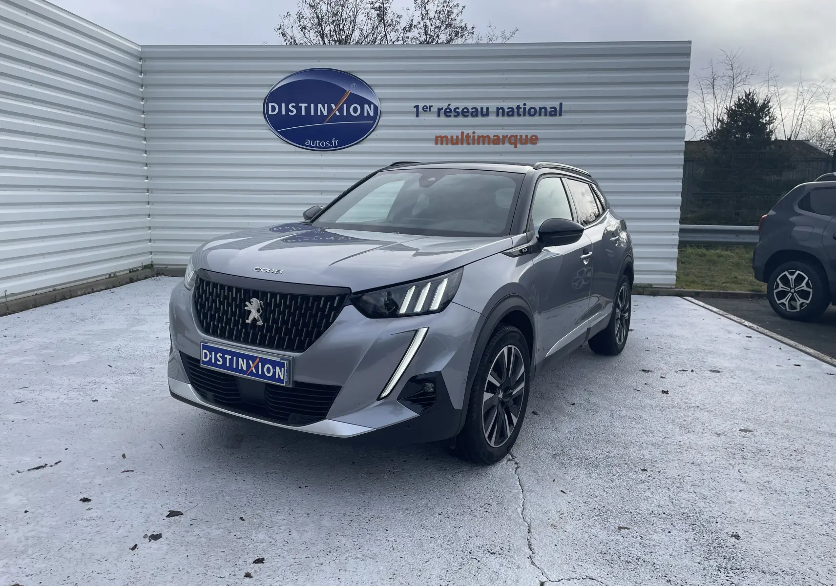 Peugeot 2008 gris Artense en 3/4 avant droit, avec calandre noire et feux LED distinctifs, stationnée devant un mur blanc.