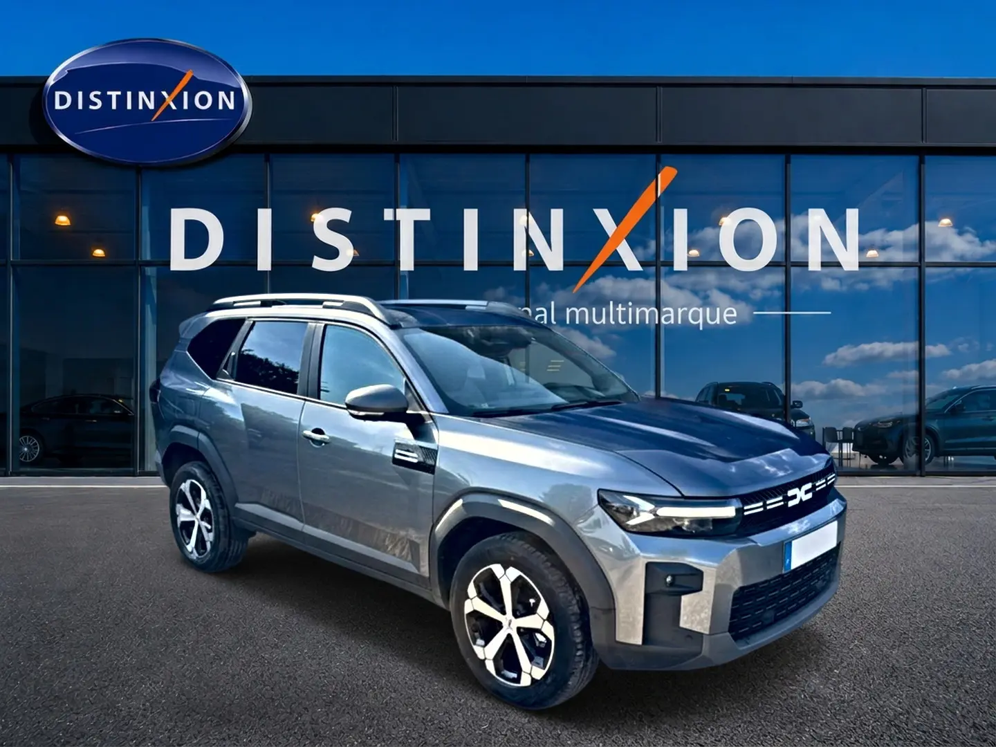Vue 3/4 avant droit d'un Dacia Bigster gris schiste avec barres de toit et jantes alu devant un showroom Distinxion.