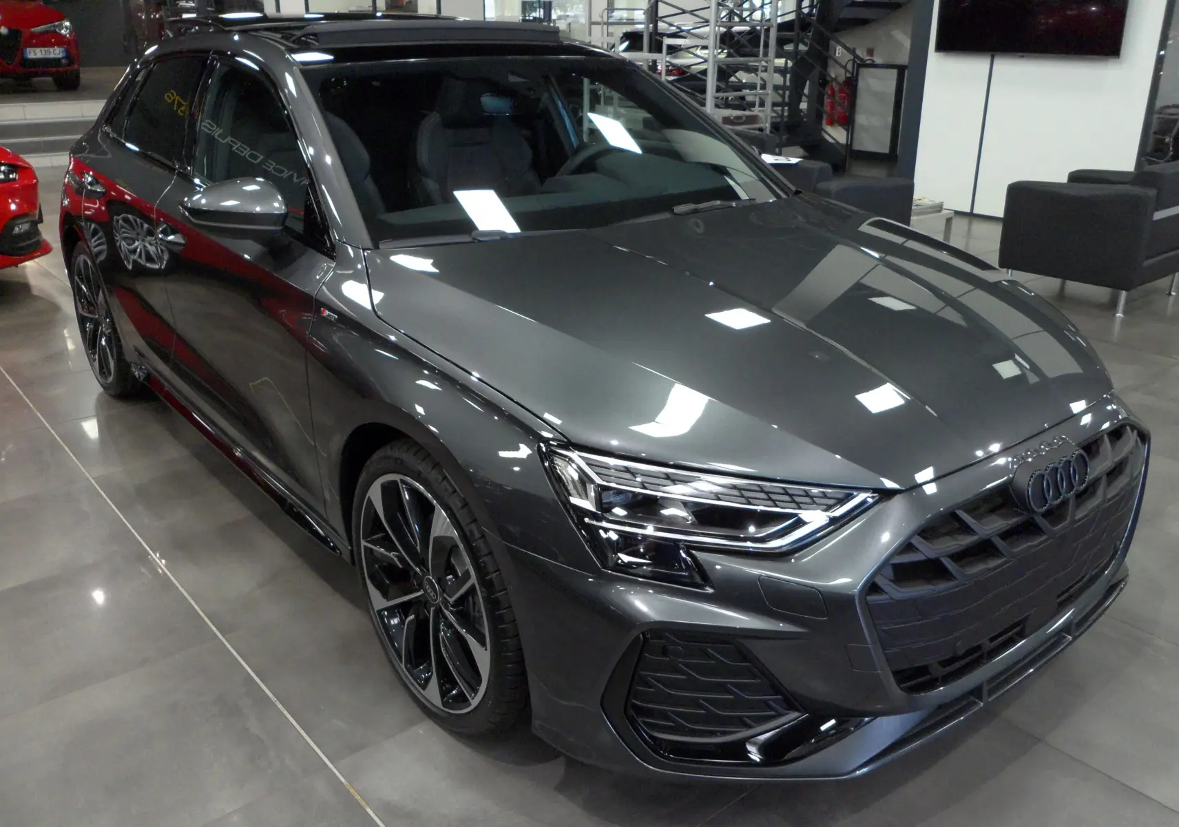 Audi A3 Sportback gris Daytona vue 3/4 avant droit en showroom, avec jantes noires bi-ton et calandre S-Line.