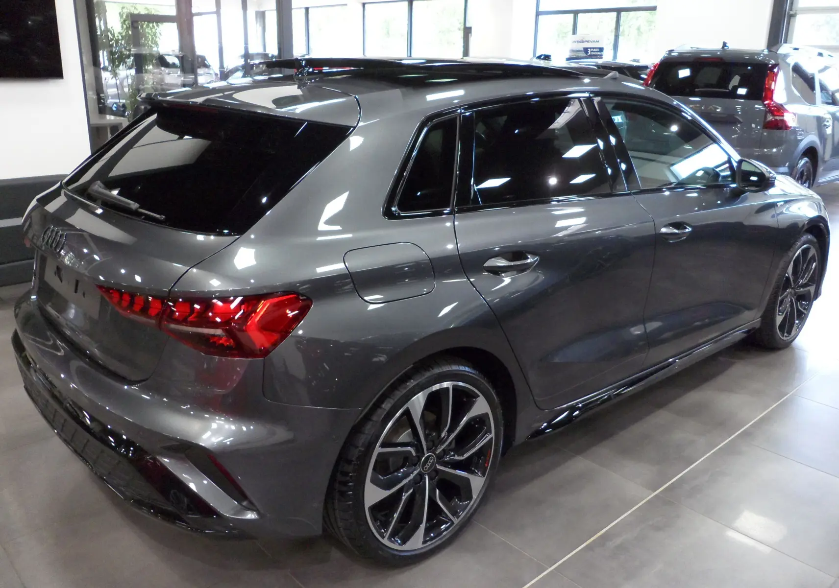 Audi A3 Sportback gris Daytona vue 3/4 arrière droit en showroom, avec jantes bi-ton et feux arrière LED allumés.