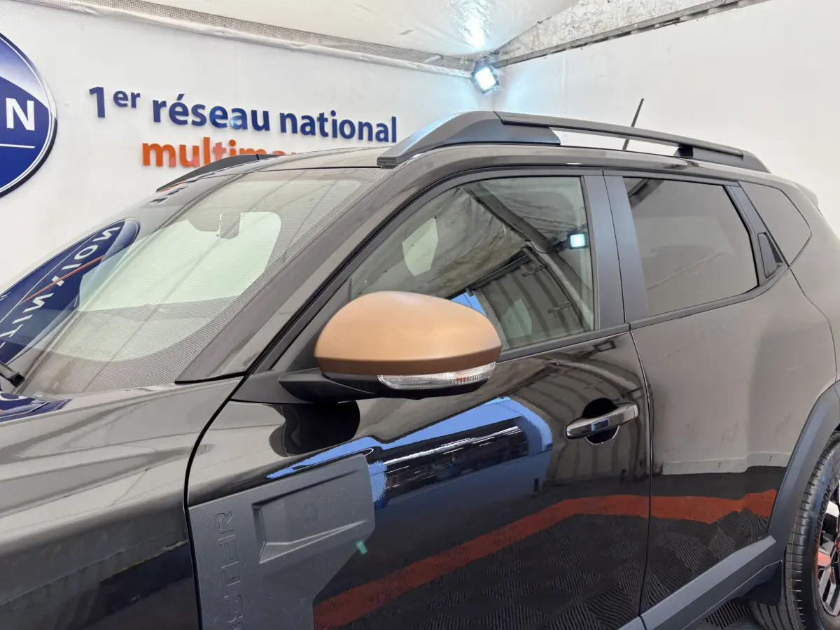 Vue latérale gauche du Dacia Duster noir avec rétroviseur couleur bronze et barres de toit noires visibles.