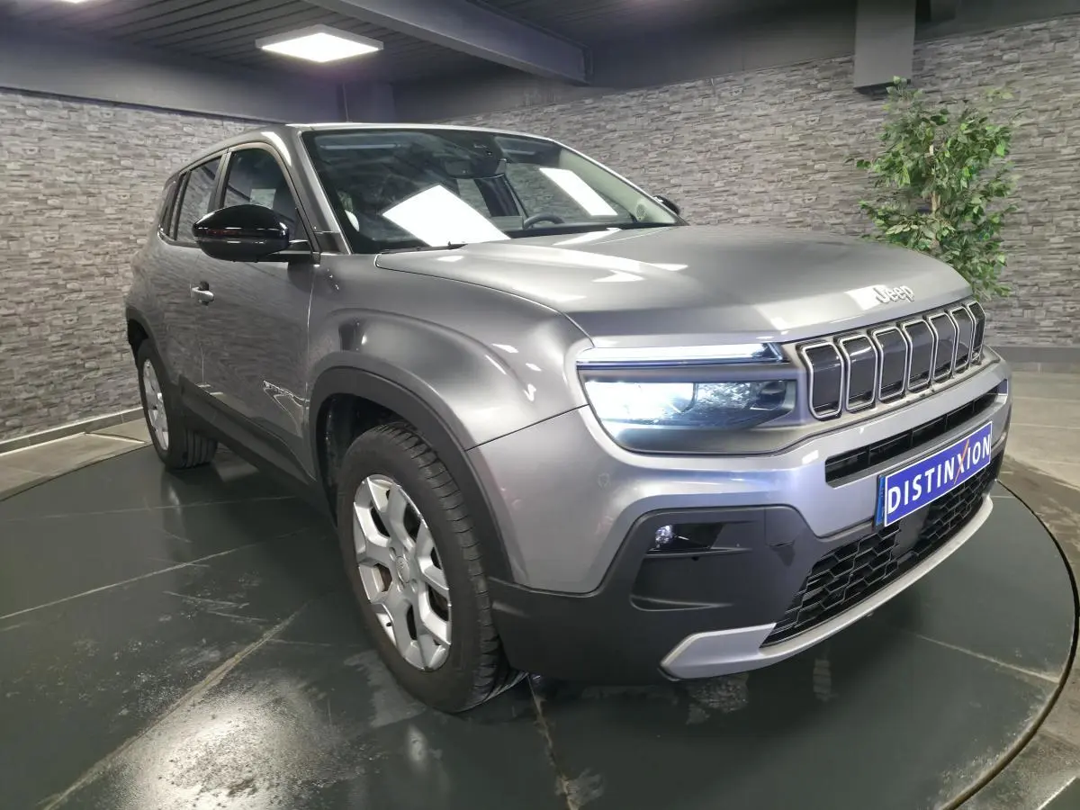 Jeep Avenger gris métallisé en 3/4 avant droit, avec calandre distinctive et phares LED allumés, en intérieur showroom.