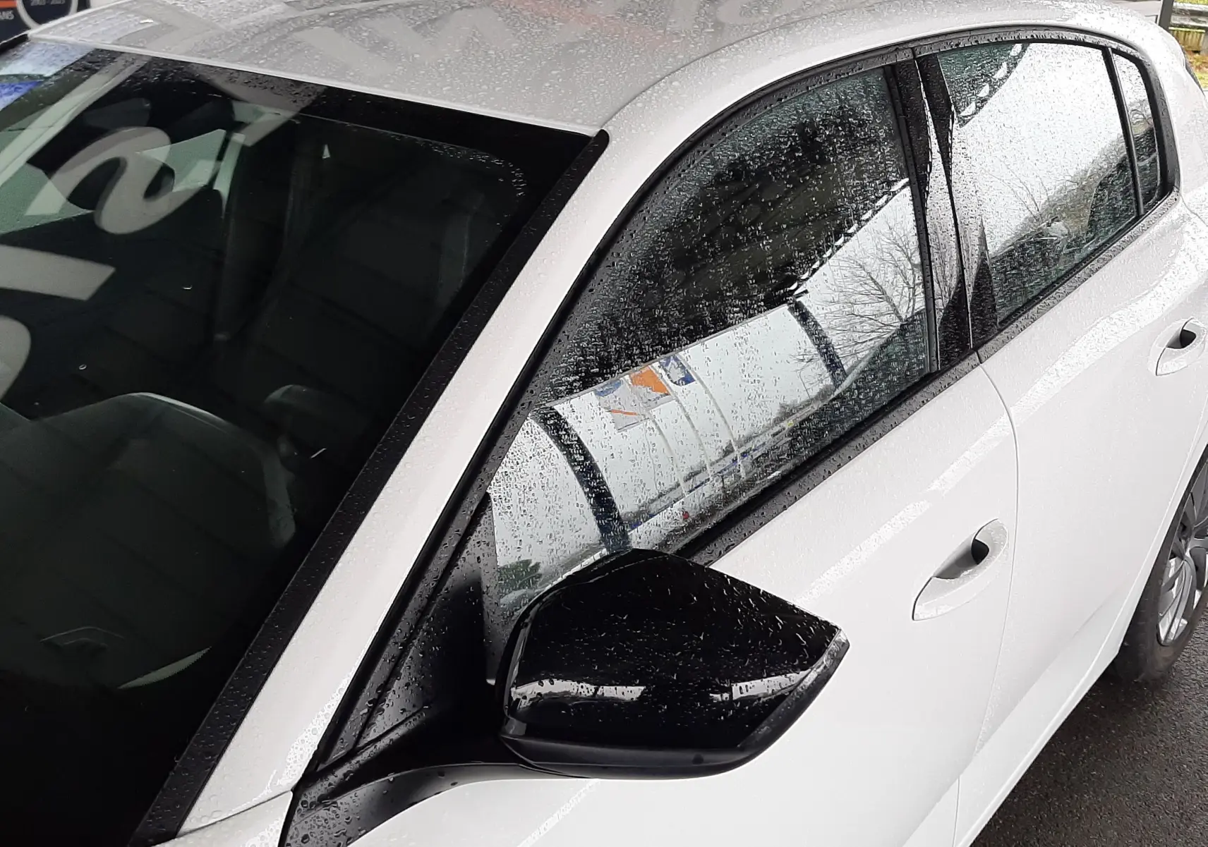 Vue côté gauche avant d'une Peugeot 308 blanche banquise avec rétroviseur noir sous la pluie, détails de la porte et vitre visibles.