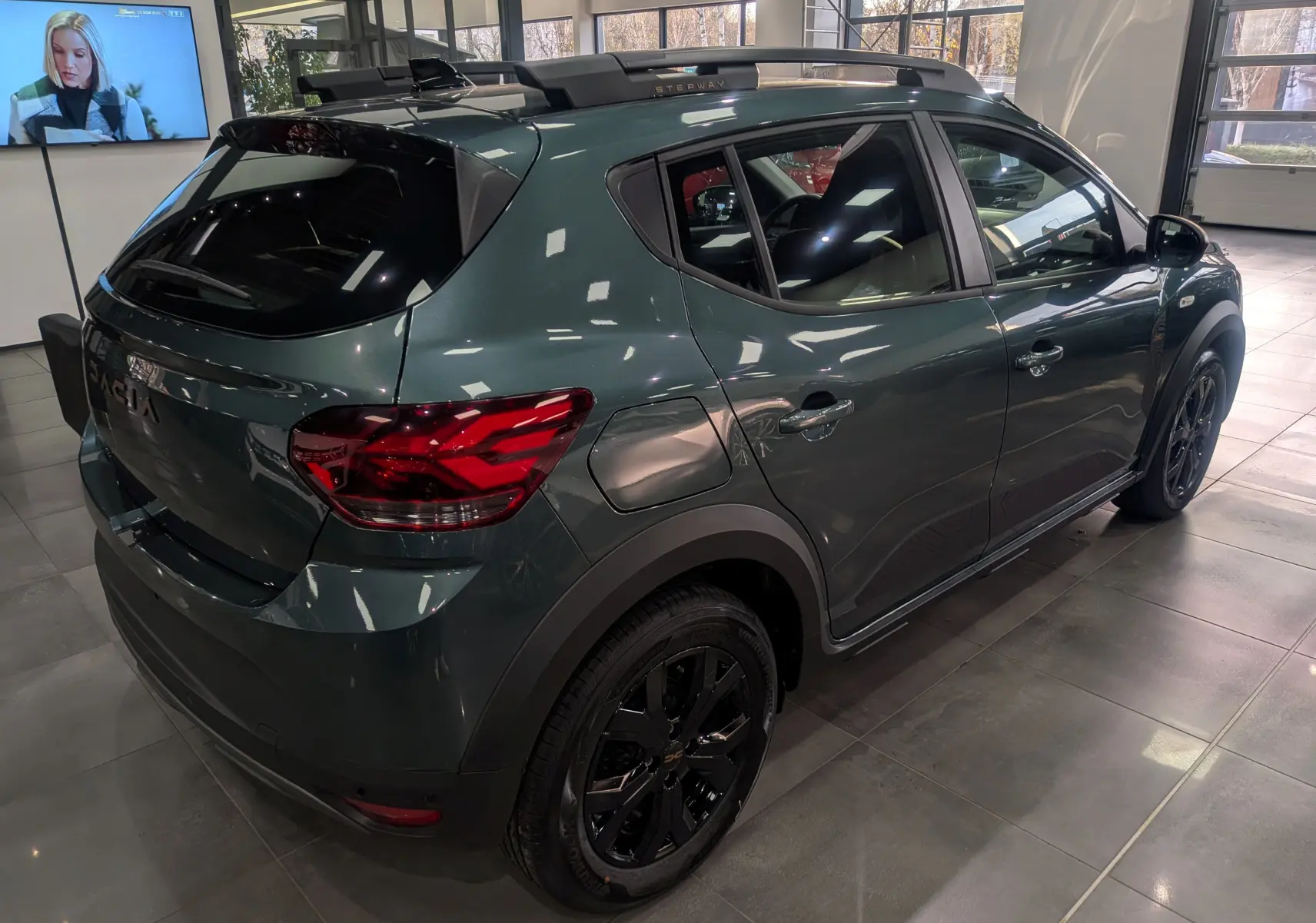 Vue 3/4 arrière droite d'une Dacia Sandero Stepway 2025 vert cèdre avec jantes noires et toit noir en showroom.