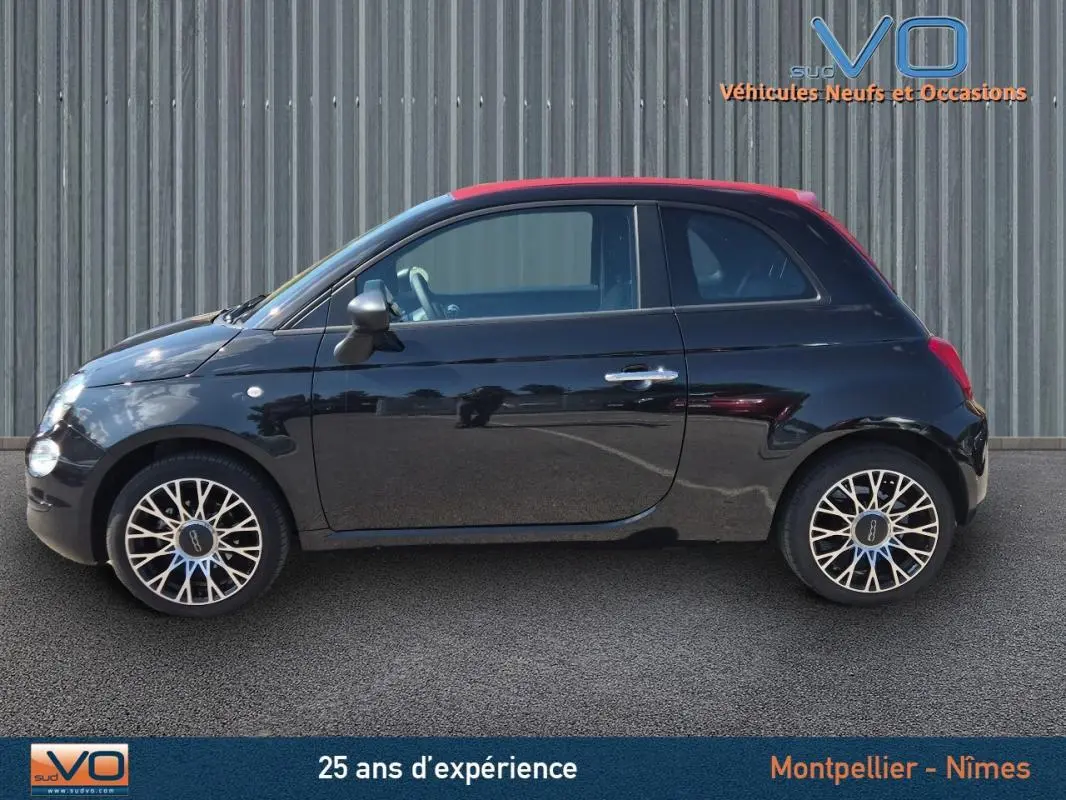 Vue de profil côté gauche d’une FIAT 500C noire 2024 avec toit rouge et jantes stylisées, sur fond industriel.