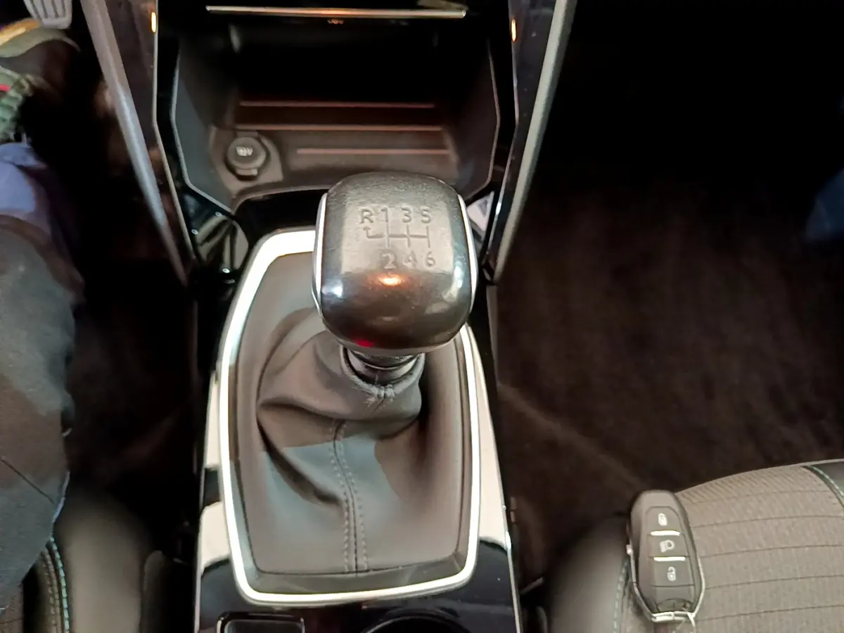 Gros plan sur le levier de vitesses manuel noir de la Peugeot 208 blanche, intérieur sombre avec siège tissu visible.