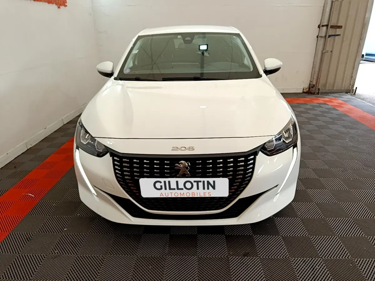 Vue frontale d'une Peugeot 208 blanche avec calandre noire et feux LED, stationnée sur un sol à damiers gris et rouge.