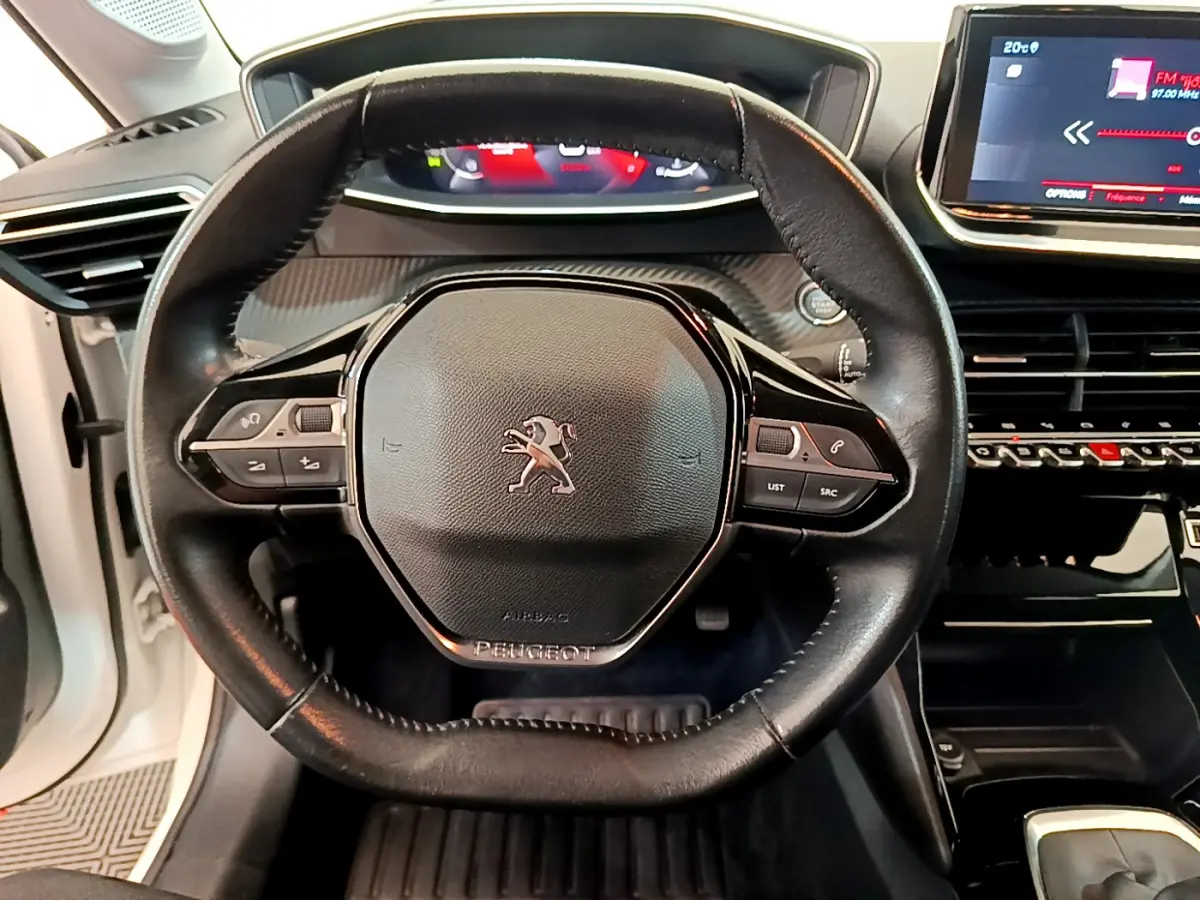 Vue rapprochée du volant cuir noir de la Peugeot 208 blanche, avec commandes intégrées et tableau de bord numérique visible.