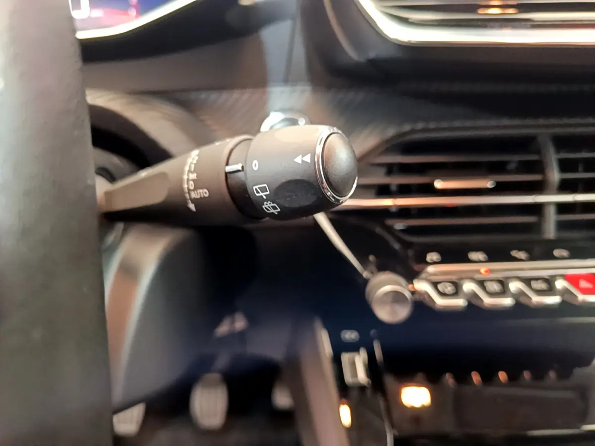 Gros plan sur la commande d'essuie-glace à gauche du volant dans l'habitacle sombre d'une Peugeot 208 blanche.