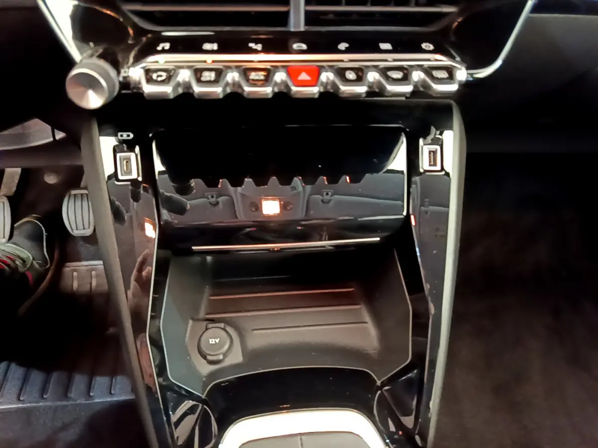 Vue rapprochée de la console centrale noire du Peugeot 208 blanc, avec prise 12V et commandes tactiles au-dessus.