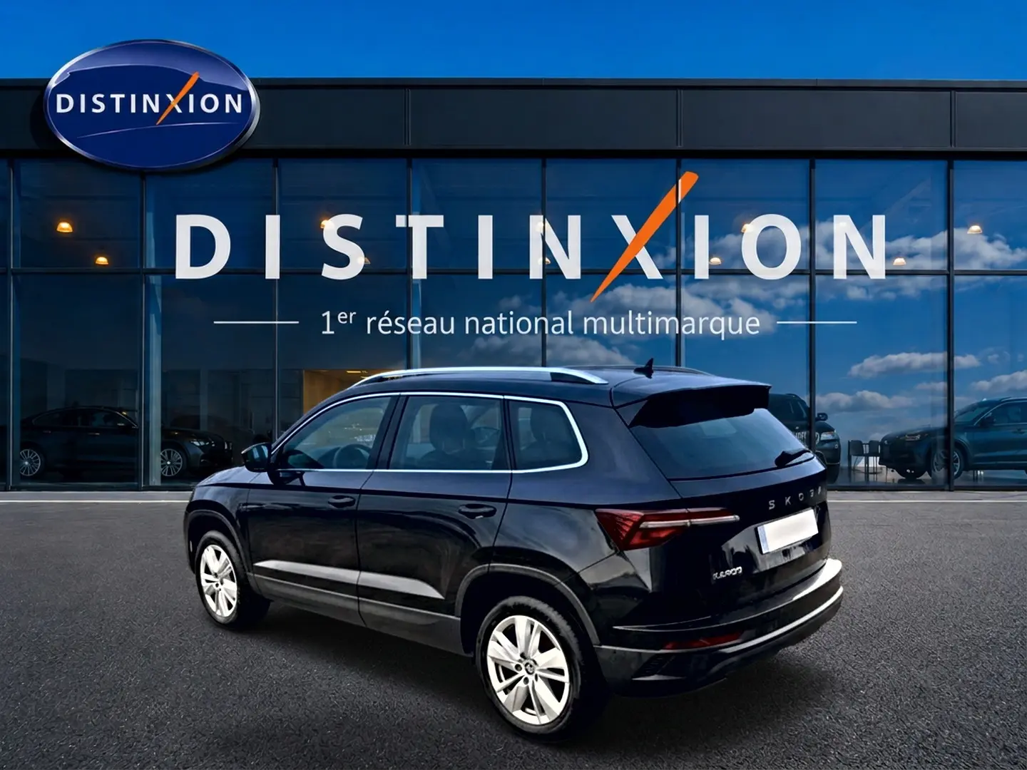 Vue 3/4 arrière droite d'un Skoda Karoq noir 2025 avec jantes alu, barres de toit et feux arrière LED, devant un showroom Distinxion.