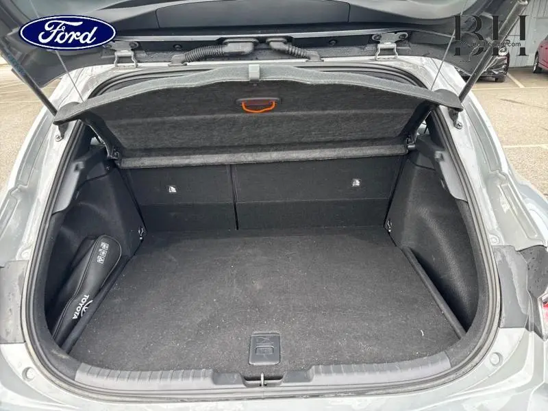 Coffre ouvert vu de l'arrière d'une Toyota Corolla 122h Dynamic Business gris Manhattan métallisé, avec tapis noir et rangement latéral.