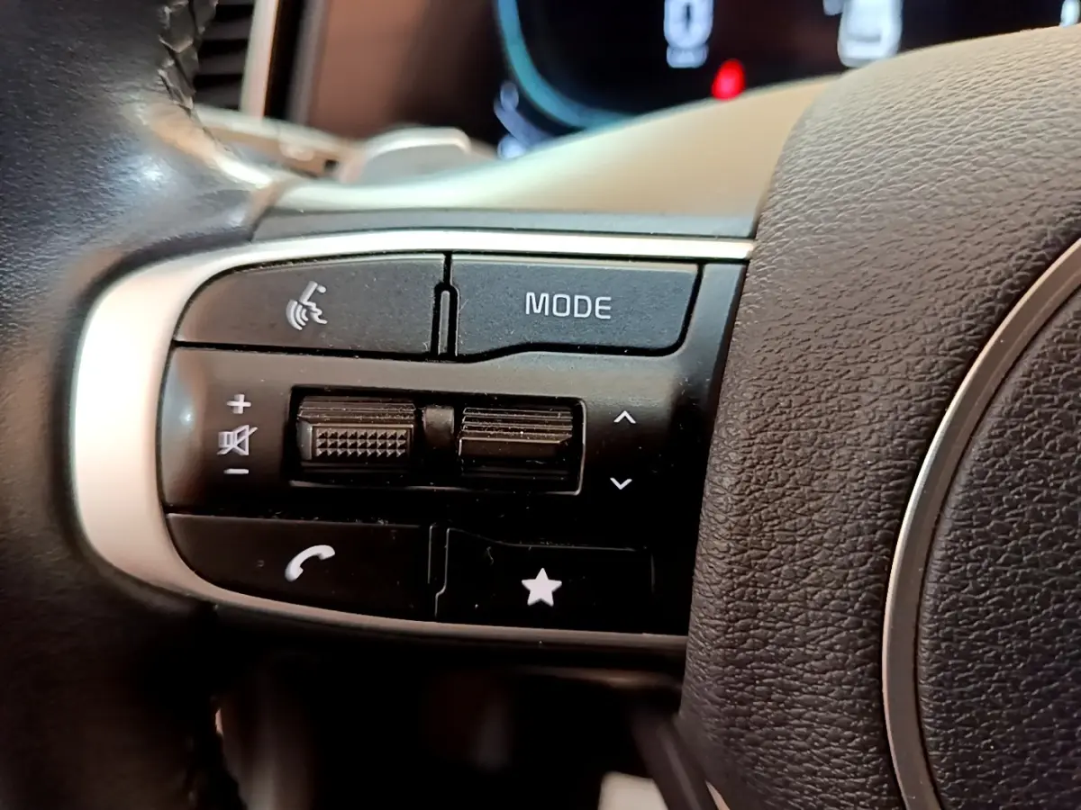 Gros plan sur les commandes gauche du volant cuir noir du Kia Sportage blanc 2022, boutons mode et téléphone visibles.