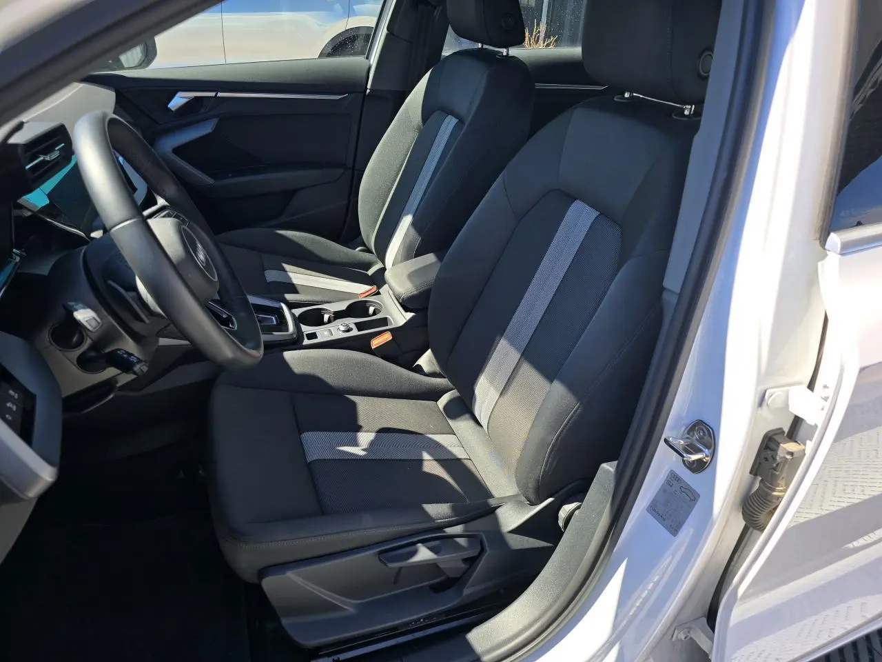 Vue intérieure côté conducteur de l'Audi A3 Sportback 2024 en Blanc Arkona, avec sièges tissu noir et console centrale visible.