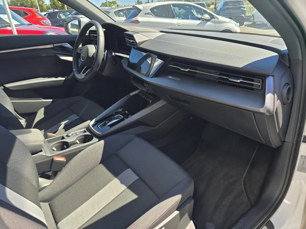 Vue intérieure côté passager d'une Audi A3 Sportback 2024 blanche, avec tableau de bord noir et sièges tissu gris foncé.