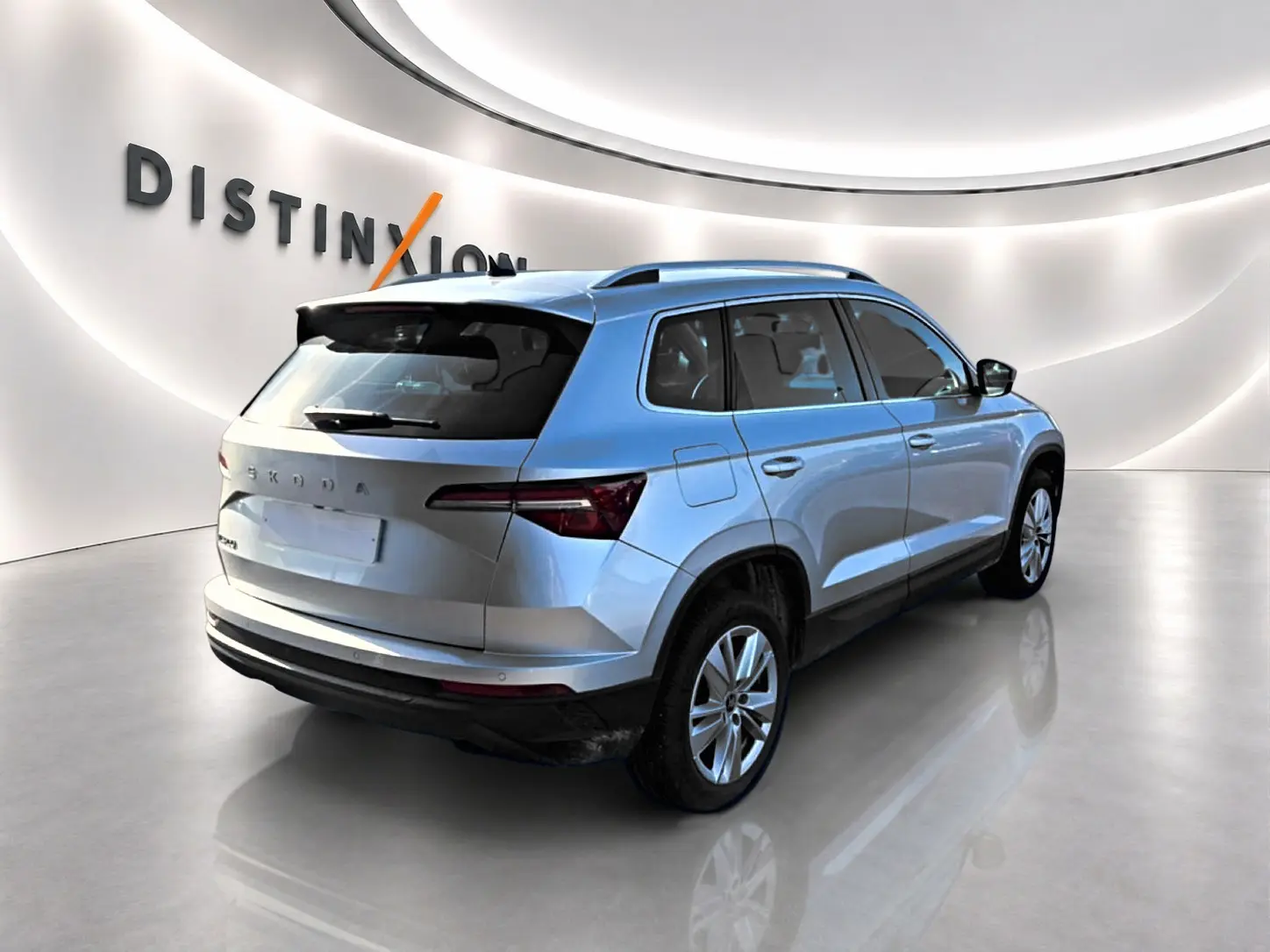 Vue 3/4 arrière droite d’un Skoda Karoq gris 2025 avec barres de toit et feux arrière LED dans un showroom lumineux.
