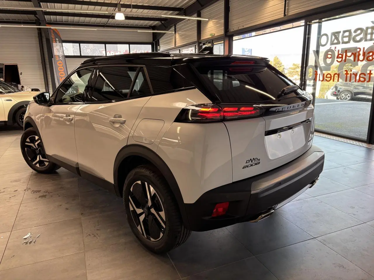 Vue 3/4 arrière droite d'un Peugeot 2008 blanc hybride essence 2025 avec toit noir et feux arrière allumés.