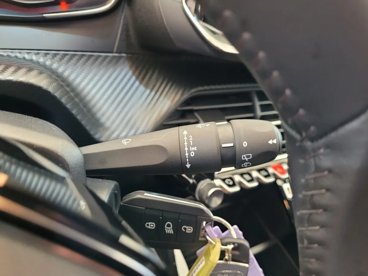 Gros plan sur la manette d'essuie-glace et clé de contact dans l'habitacle d'une Peugeot 208 gris foncé, volant cuir visible.
