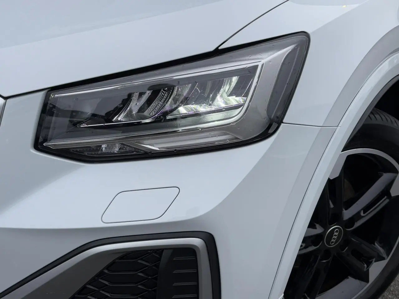 Gros plan sur le phare avant droit et la jante noire polie de l'Audi Q2 blanc glacier 30 TFSI S-Line 2024.