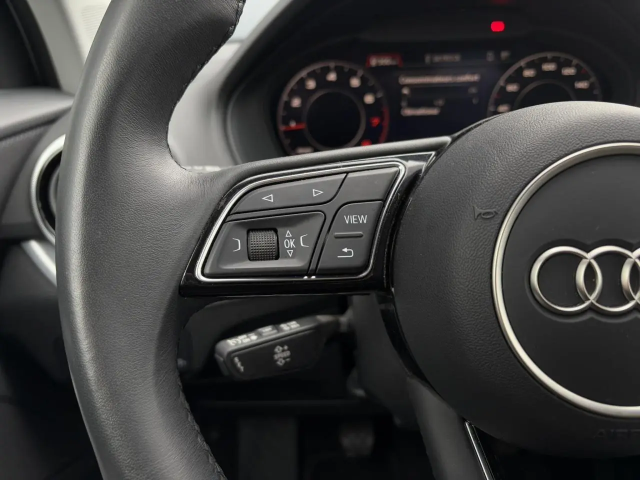 Gros plan sur le volant cuir noir multifonction gauche de l’Audi Q2 30 TFSI 110 S-Line avec affichage numérique en arrière-plan.