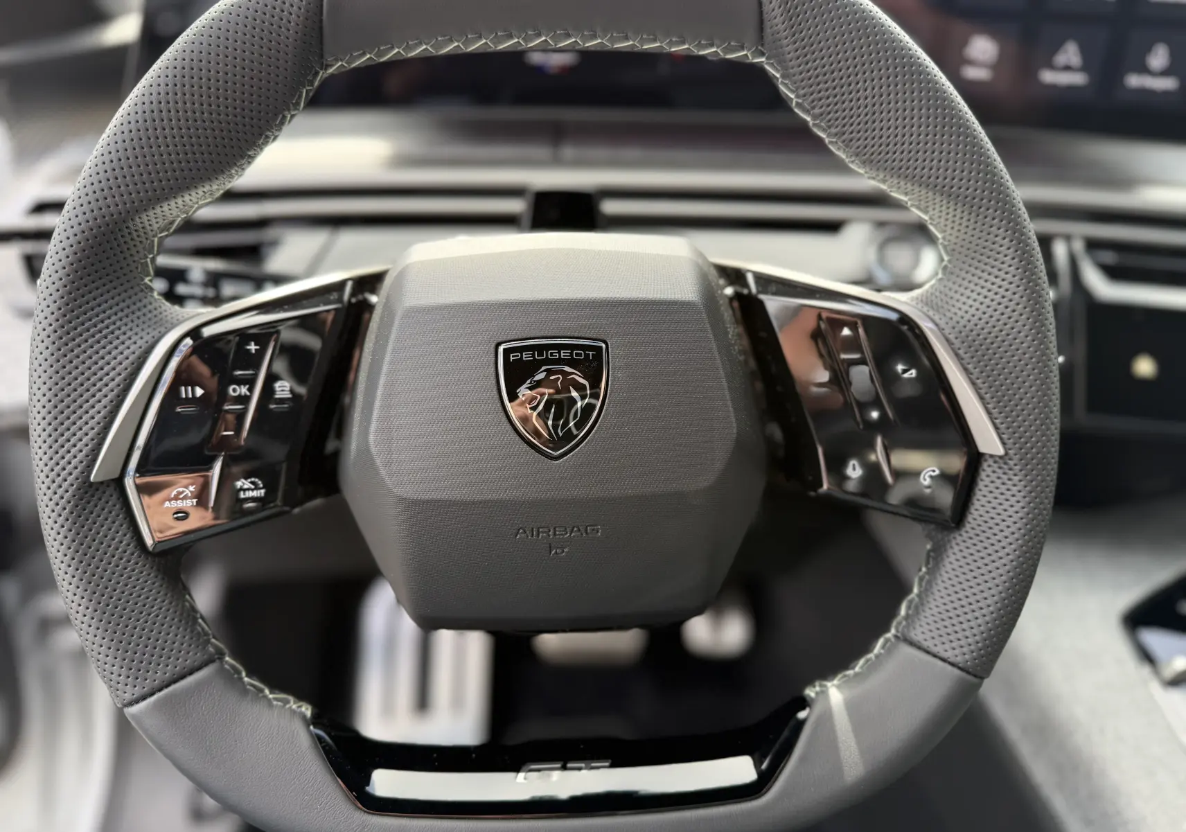 Volant en cuir perforé gris avec commandes intégrées et logo Peugeot au centre, vue frontale rapprochée.
