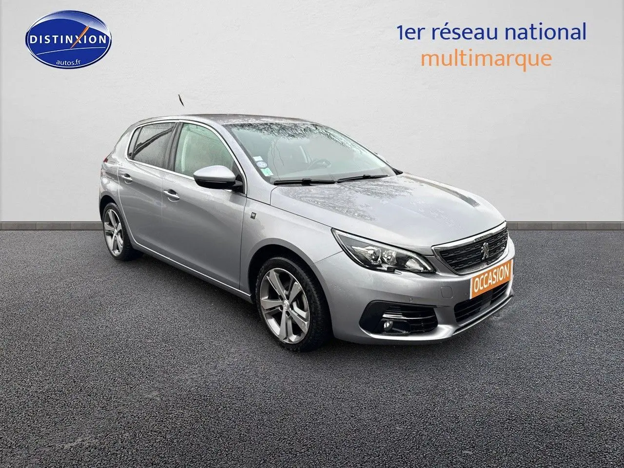 Peugeot 308 gris Artense vue 3/4 avant droit sur sol gris avec plaque orange "OCCASION" visible.