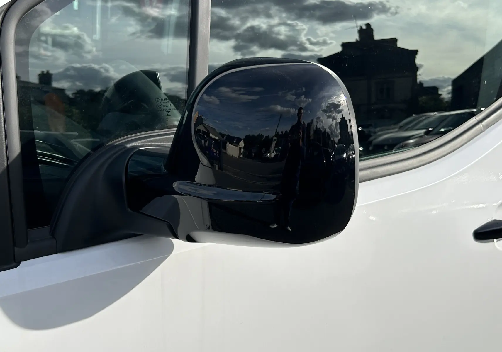 Rétroviseur noir en gros plan côté gauche d'un Peugeot Partner Fourgon blanc sous un ciel nuageux.