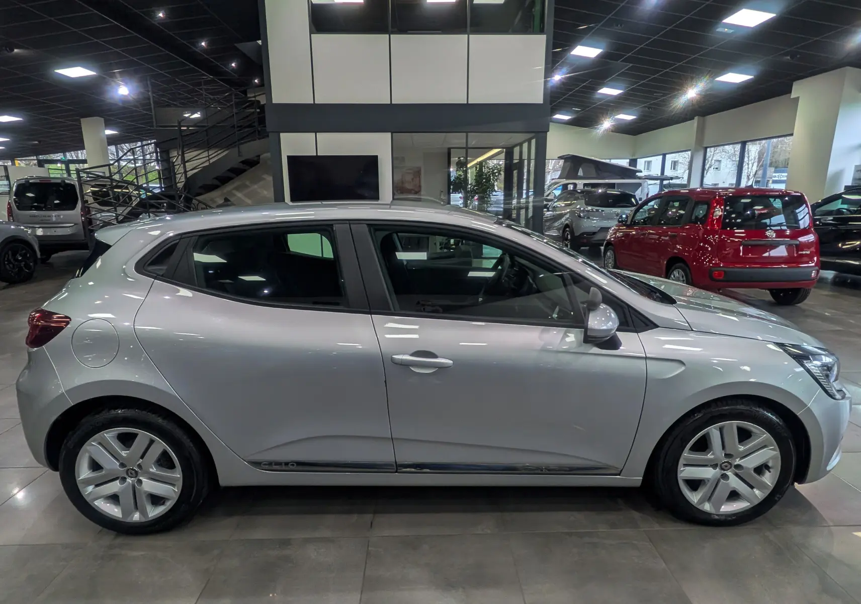 Profil côté gauche d'une Renault Clio V gris platine en intérieur, avec jantes alliage et lignes épurées visibles.