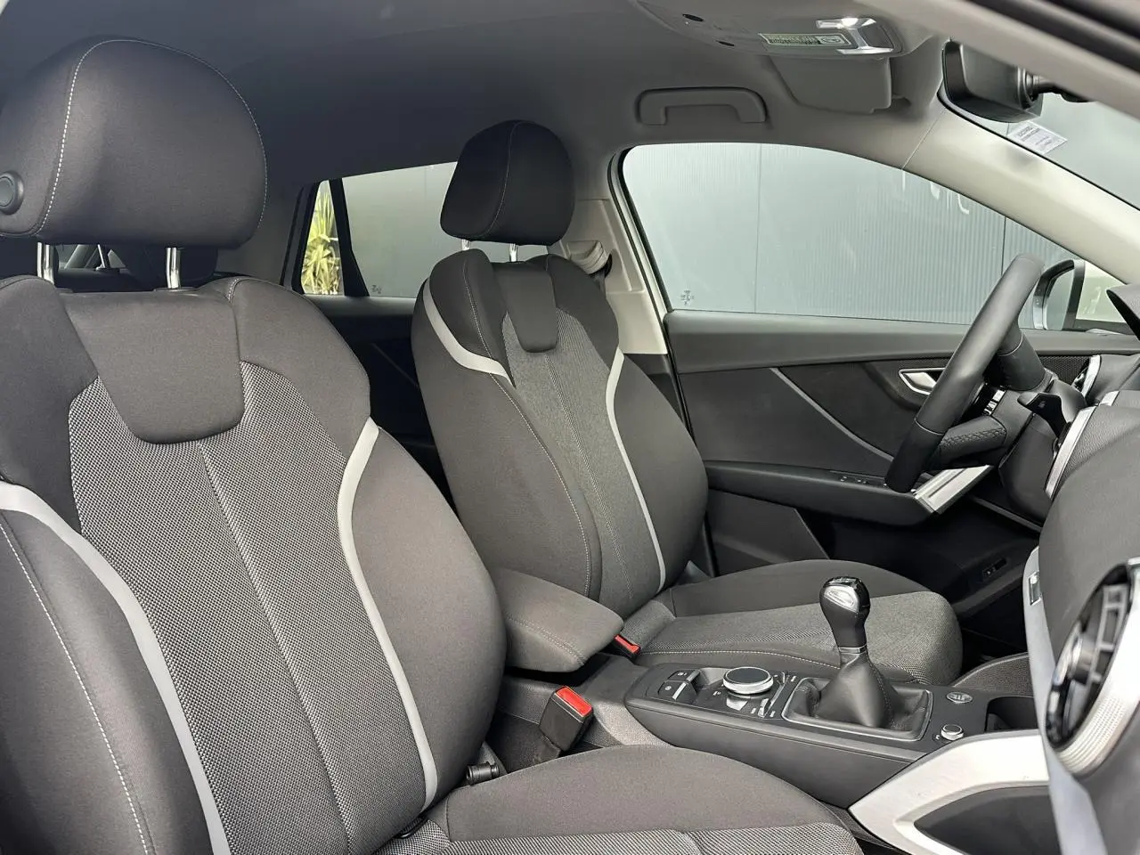 Vue intérieure côté conducteur et passager avant de l'Audi Q2 30 TFSI S-Line 2024 avec sièges sport tissu gris et levier de vitesses manuel.