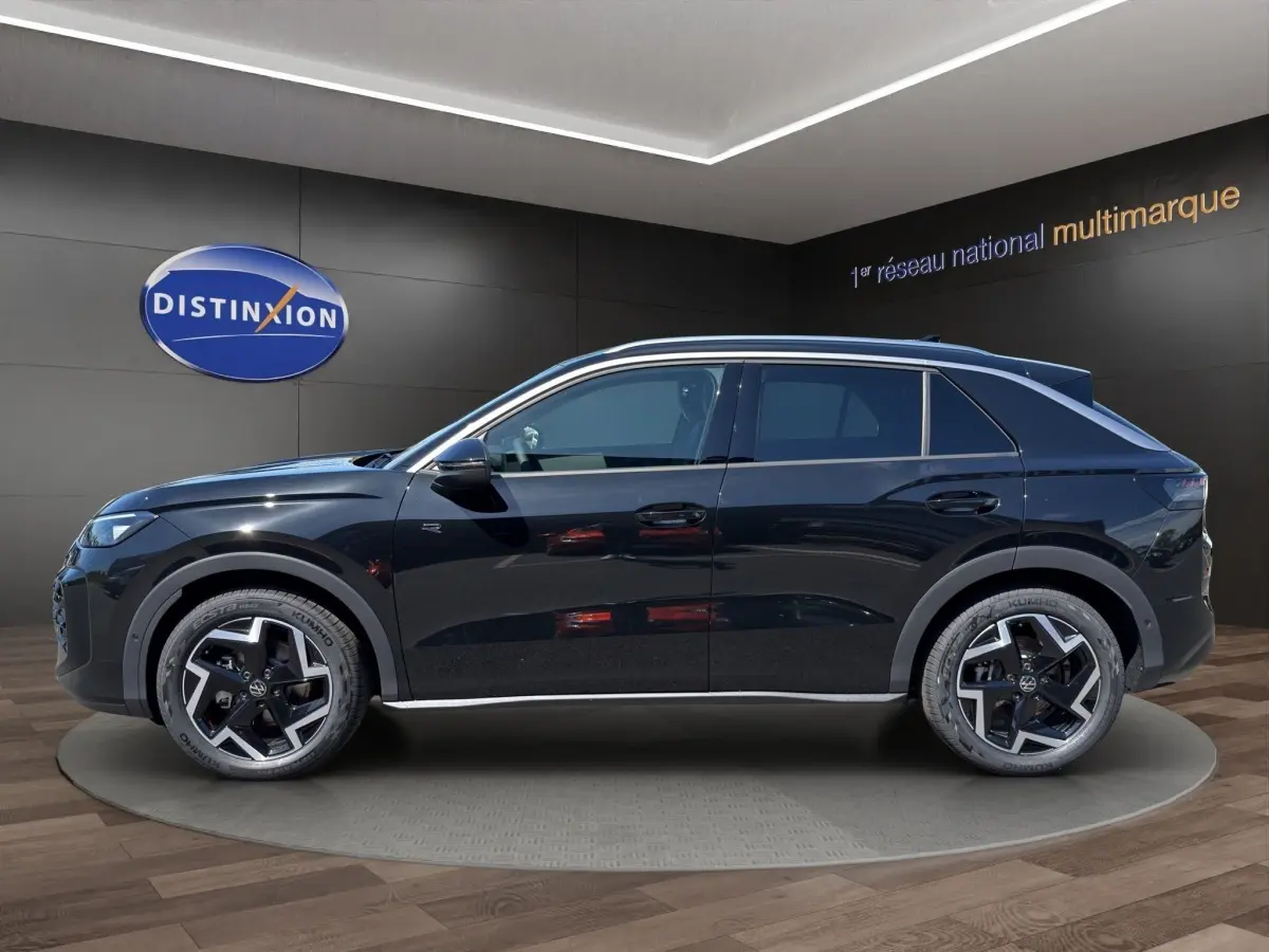 Vue latérale gauche d'un Volkswagen T-Roc noir 2026 avec jantes distinctives et toit chromé dans un showroom.