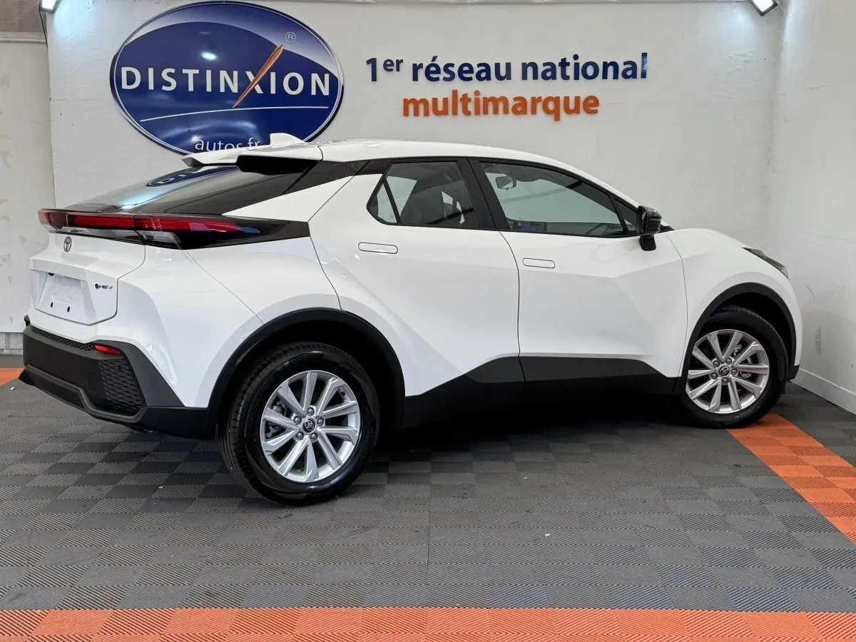 Profil droit du Toyota C-HR blanc 1.8 Hybride 140 Dynamic 2024 avec toit noir et jantes alliage visibles en intérieur showroom