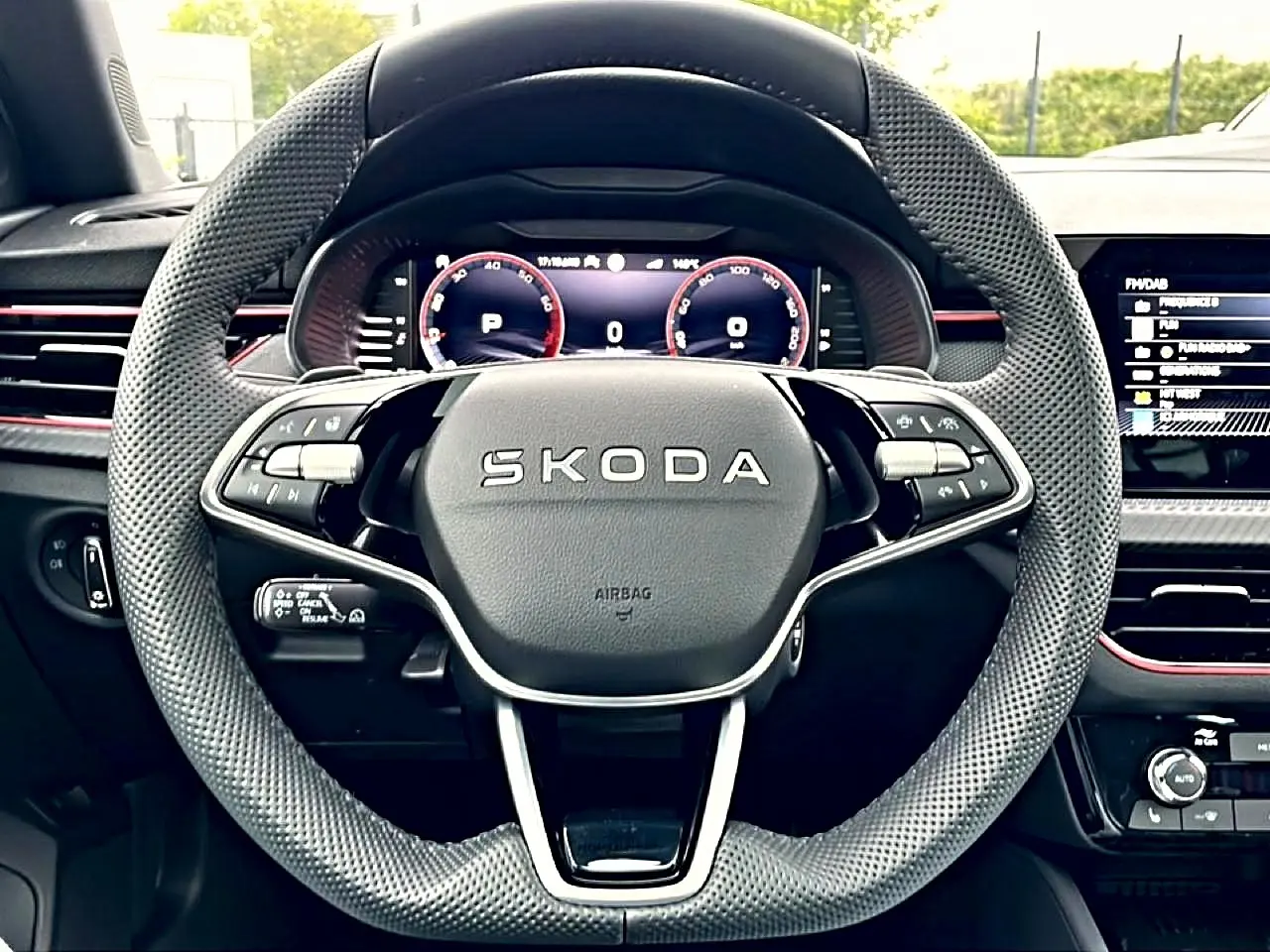 Volant sport en cuir perforé avec logo SKODA, tableau de bord numérique et détails rouges intérieurs visibles de face.