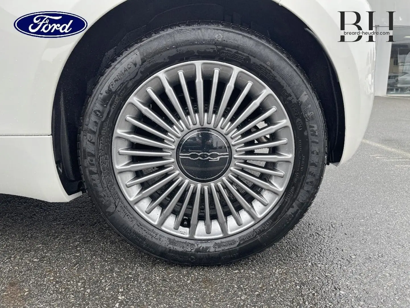 Gros plan sur la jante alu argentée d'une FIAT 500 Ice White pastel, roue avant droite visible sur sol humide.
