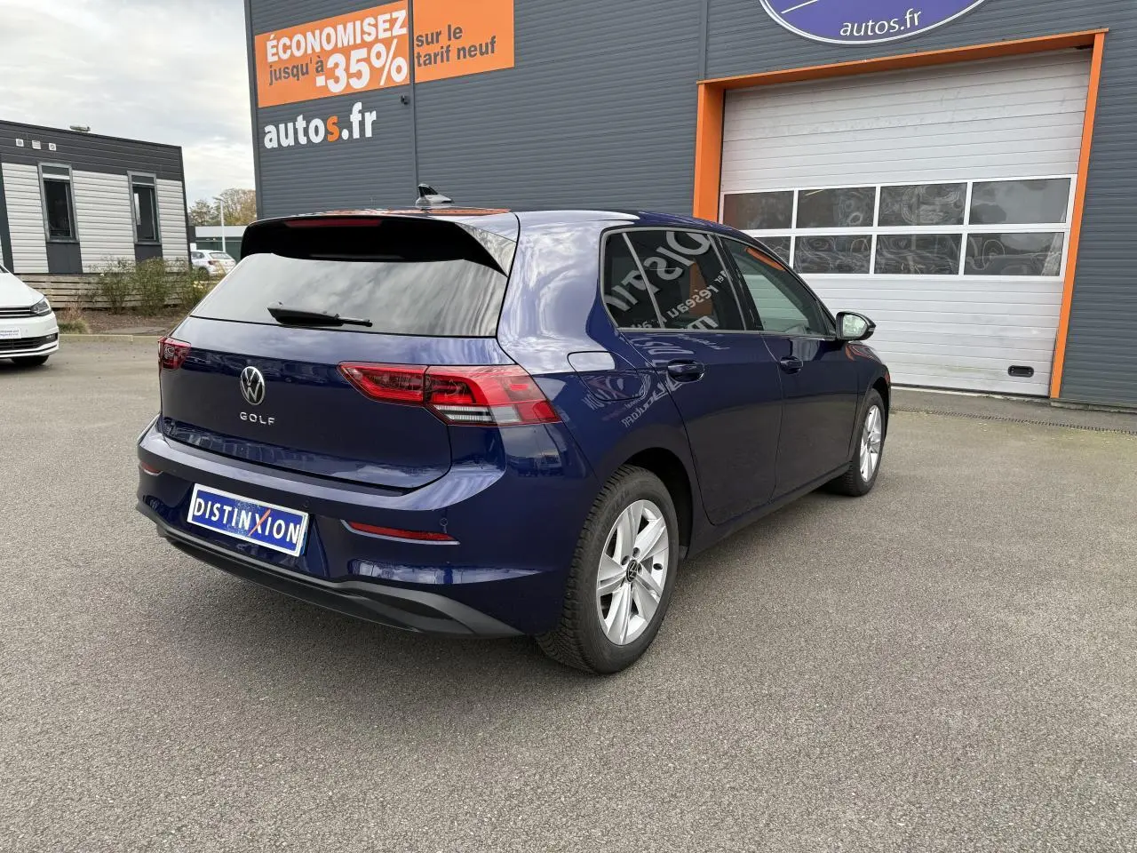 Vue 3/4 arrière droite d'une Volkswagen Golf bleu Atlantic métallisée avec vitres arrière surteintées, stationnée devant un garage autos.fr