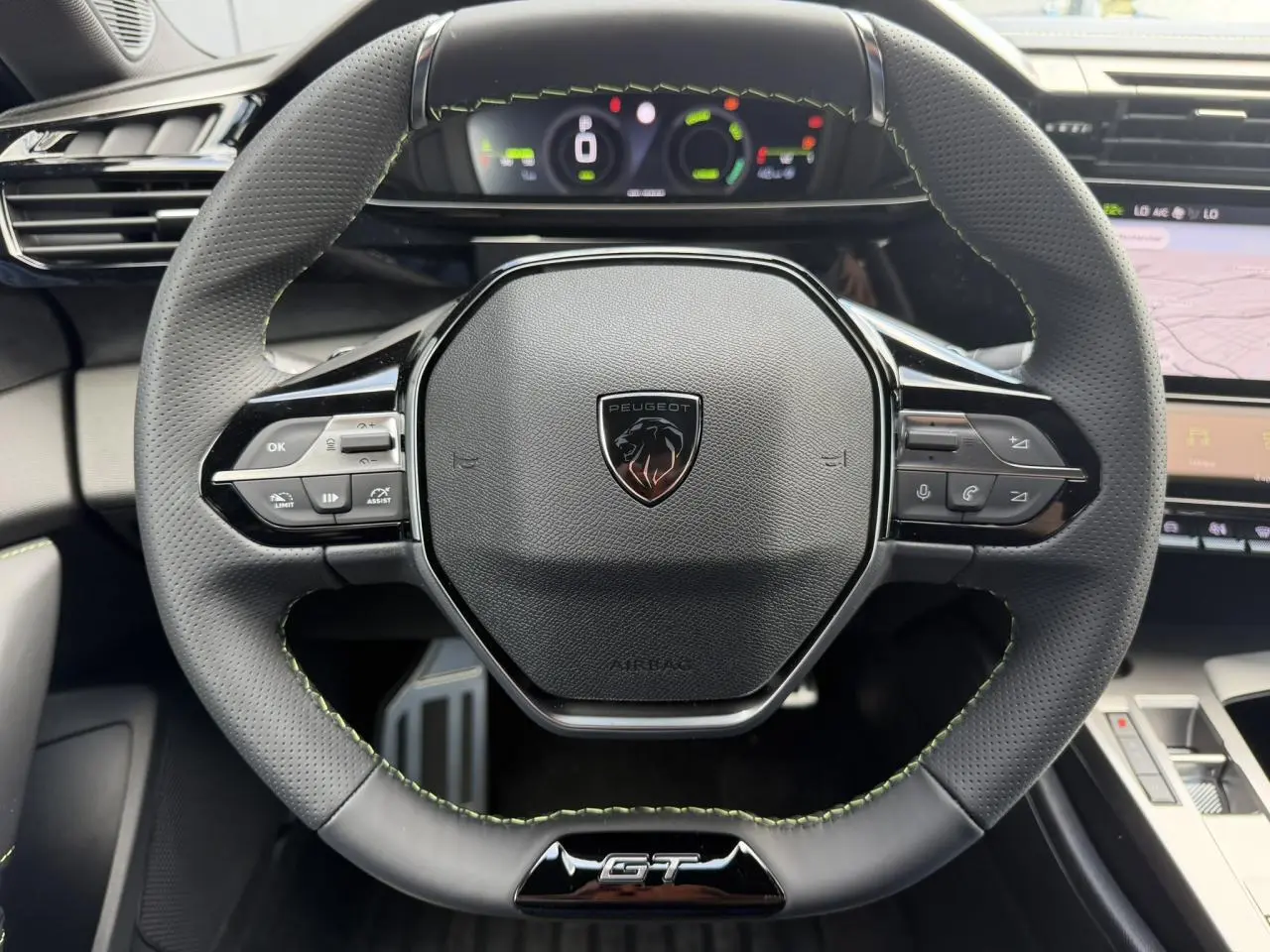 Volant compact GT en cuir noir avec surpiqûres vert Adamite et logo Peugeot, tableau de bord numérique en arrière-plan.