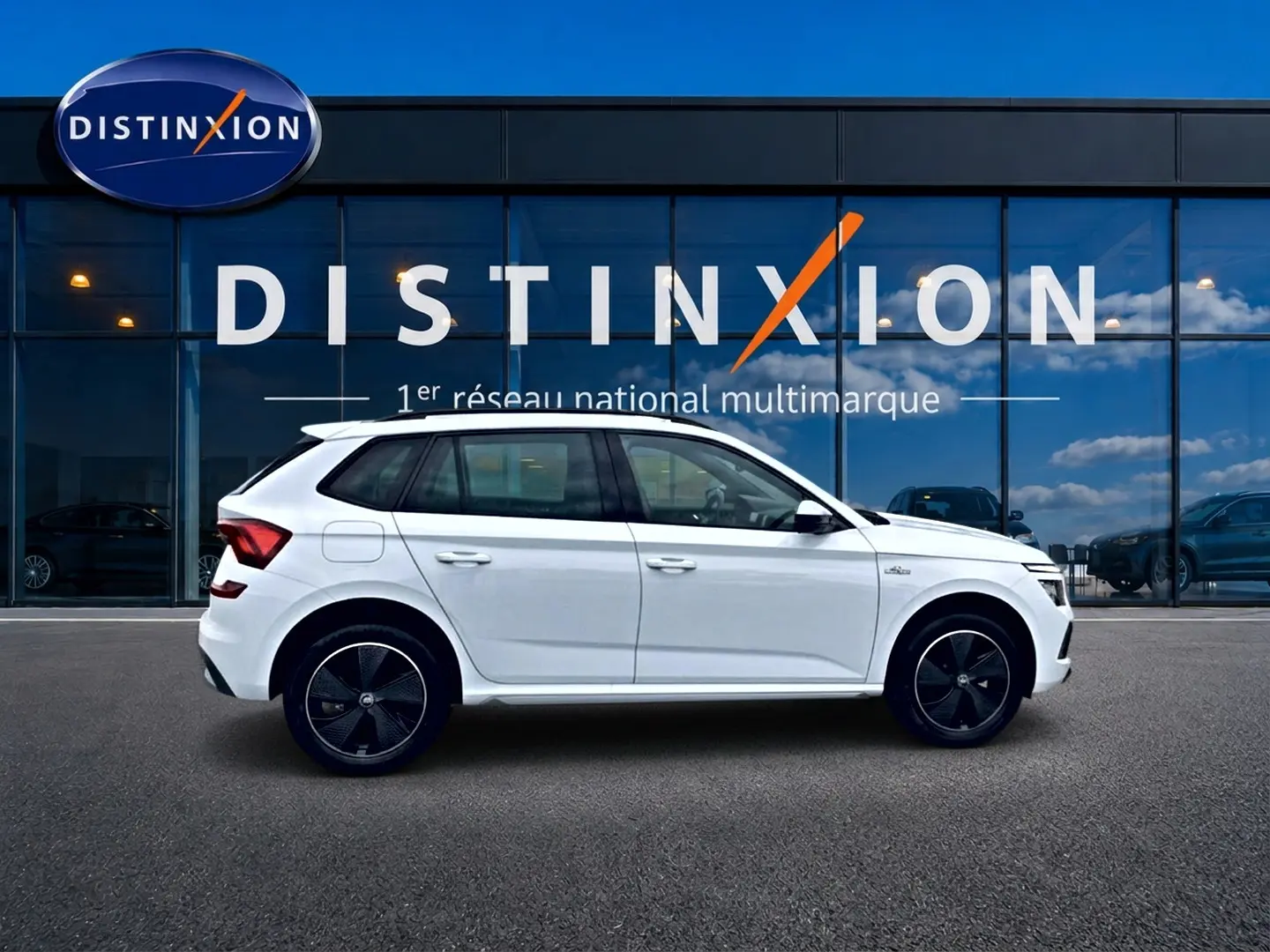 Vue latérale droite d'un Skoda Kamiq blanc nacré avec jantes noires devant un bâtiment vitré Distinxion.