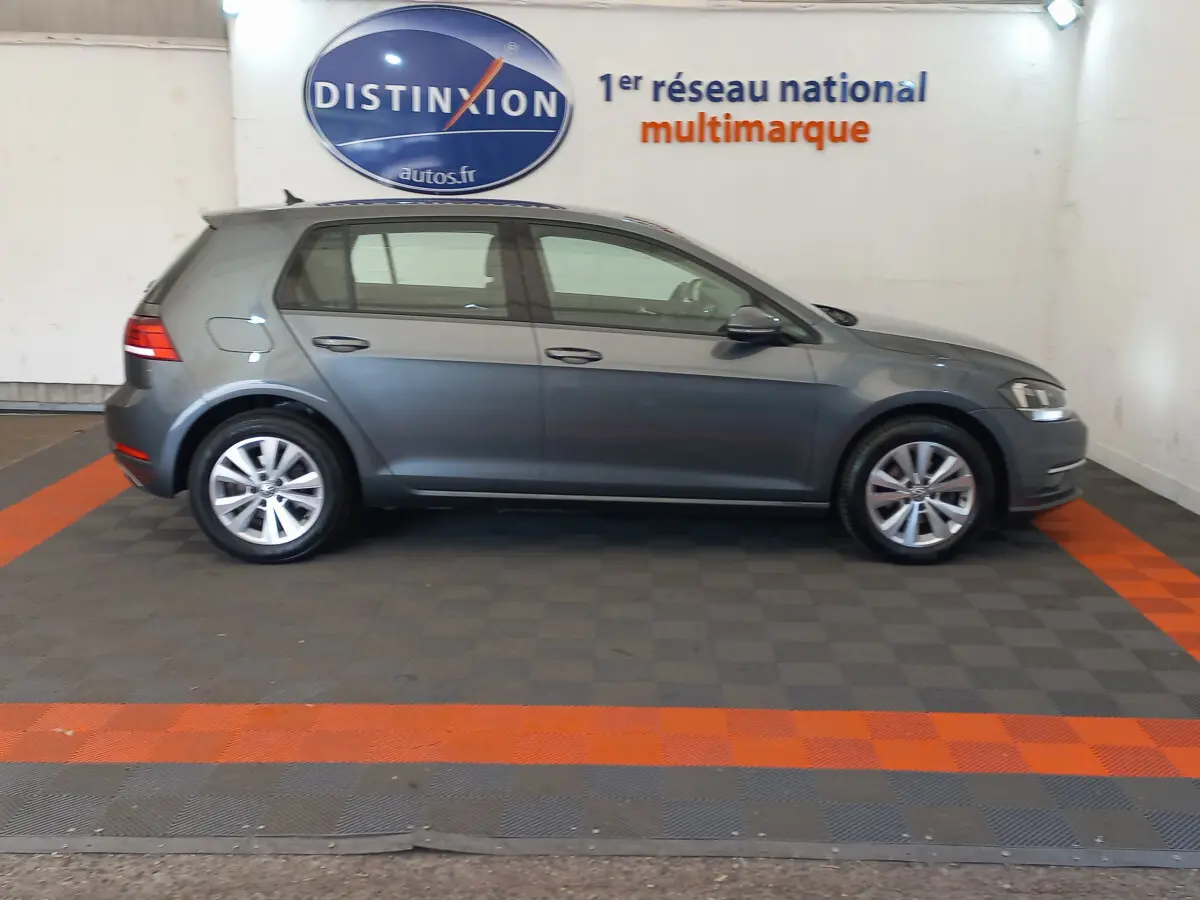 Profil côté droit d'une Volkswagen Golf gris foncé 2019 garée en intérieur sur sol à damiers avec logo Distinxion en arrière-plan.