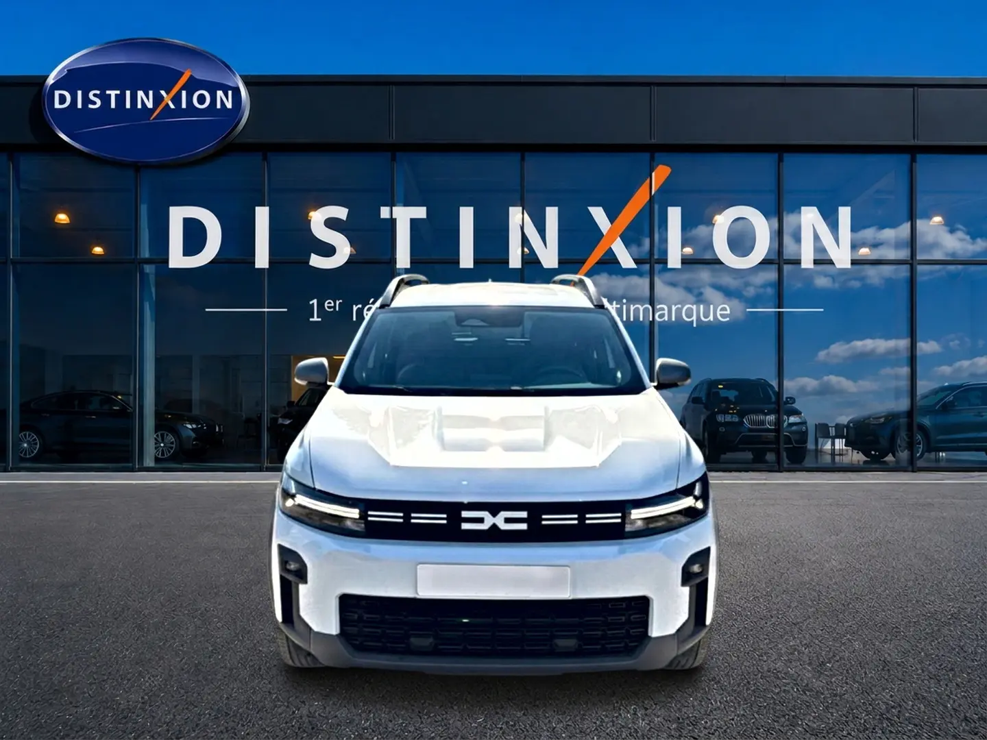 Dacia Bigster blanc vue de face, avec calandre noire distinctive et barres de toit visibles, devant une vitrine Distinxion.