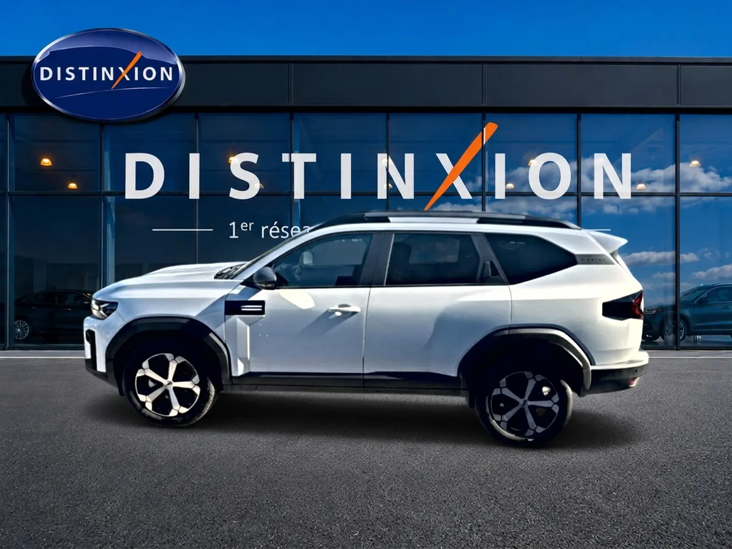 Vue de profil côté gauche d’un Dacia Bigster blanc 2026 avec barres de toit et jantes alu devant une vitrine Distinxion.