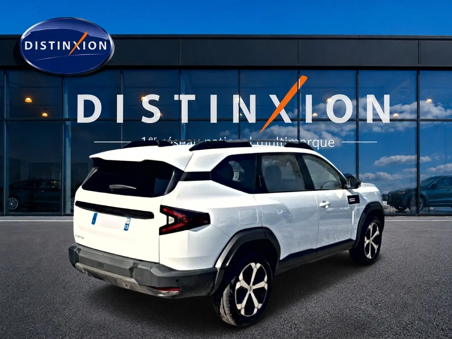 Vue 3/4 arrière droite du SUV blanc Dacia Bigster 2026 avec jantes alu et toit noir devant un showroom Distinxion.