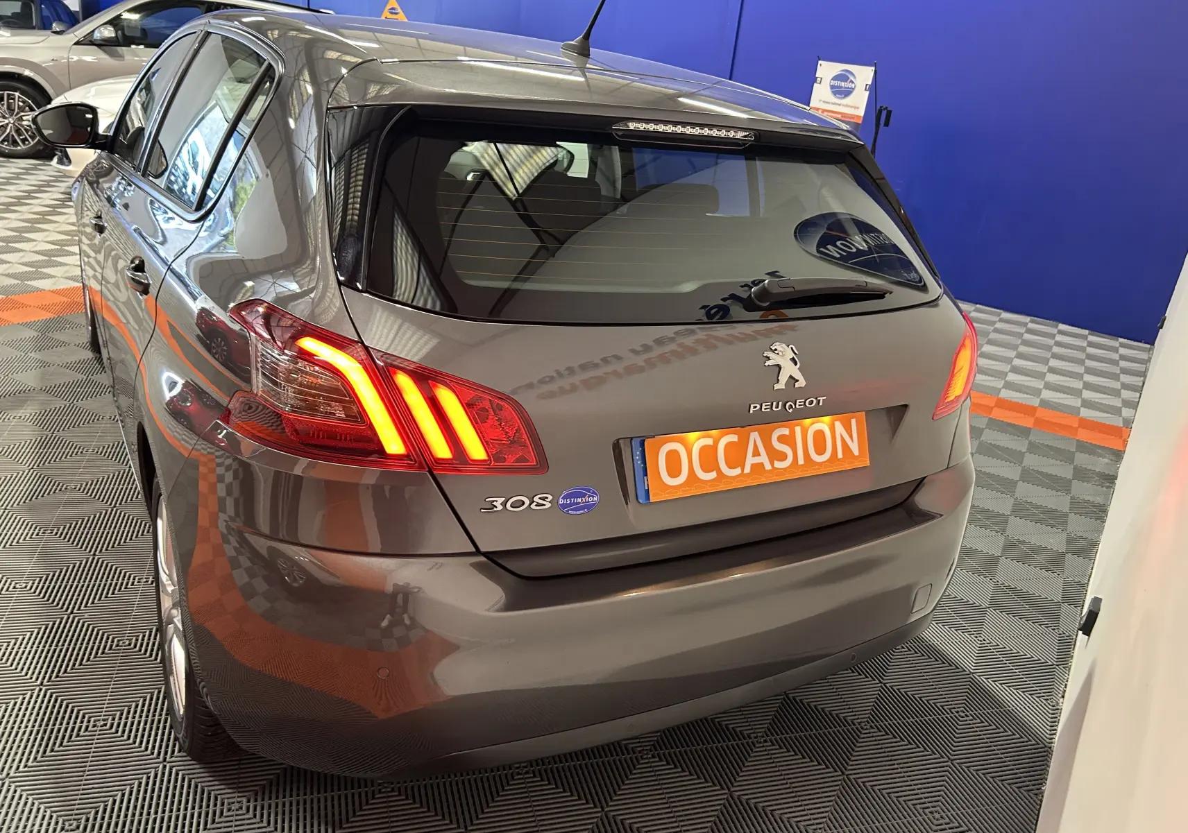 Vue 3/4 arrière droite d'une Peugeot 308 gris métallisé avec feux arrière allumés dans un showroom.