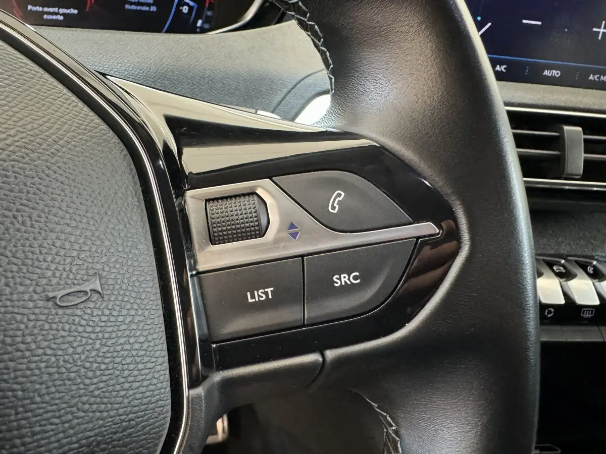 Gros plan sur les commandes gauche du volant cuir noir du Peugeot 3008 blanc, avec tableau de bord et écran tactile en arrière-plan.