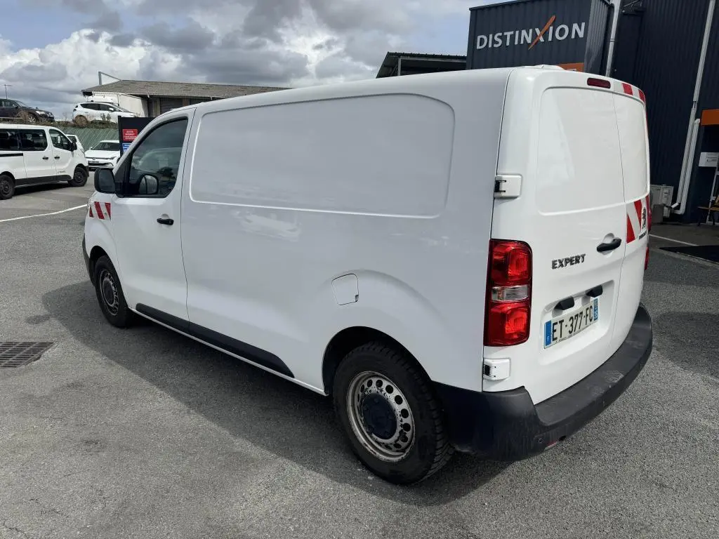 Vue 3/4 arrière droit d'un Peugeot Expert Fourgon blanc avec boucliers noirs et marquages rouges sur l'aile avant.