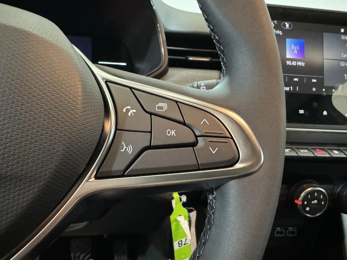 Gros plan sur les commandes gauche du volant cuir noir de la Renault Clio gris clair 2025 avec écran tactile en arrière-plan.