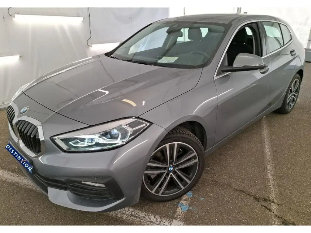 BMW Série 1 gris vue 3/4 avant droit, phares allumés et jantes alliage distinctives, stationnée en intérieur.